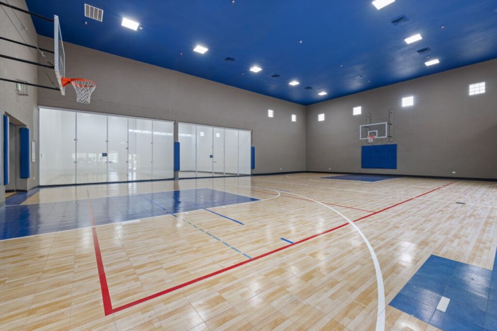Indoor basketball court with polished wooden floor, blue and red court markings, two basketball hoops, high ceiling, and large glass doors along one wall. The space is well-lit with several small windows.
