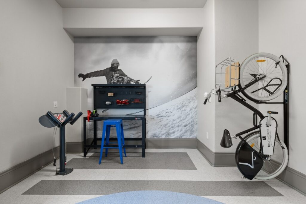 A room with a mural of a snowboarder on the back wall, a blue stool at a black workbench, a magazine stand with books, and two bicycles mounted vertically on a rack against a white wall.