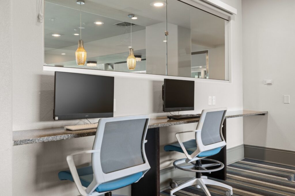 Two modern office chairs are placed at a long desk with two desktop computers, set against a white wall with a large window above. Warm pendant lights hang in the background.