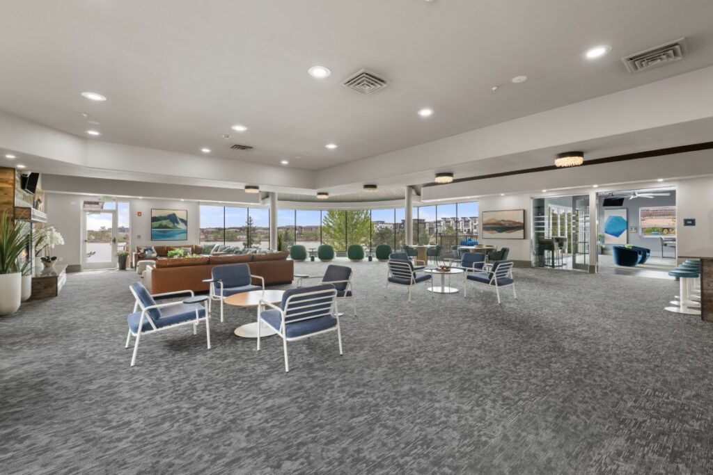 Spacious lounge with modern furniture, blue chairs, round tables, brown couch, large windows, and outdoor view. Neutral decor and bright lighting create a welcoming atmosphere.