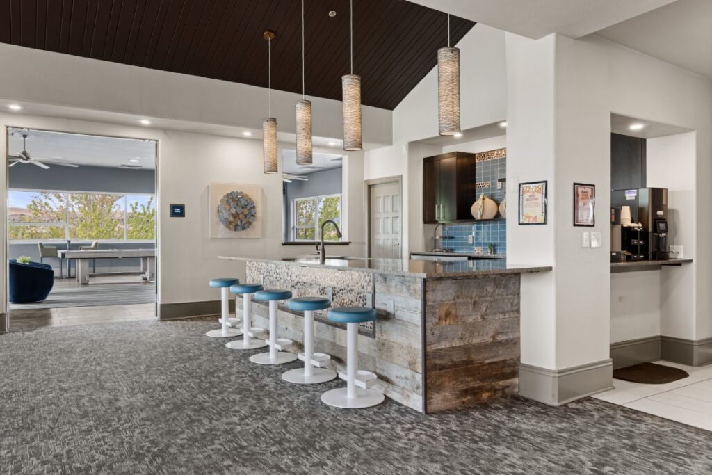 Modern kitchen with a rustic wood bar, blue stools, pendant lights, and a coffee station. Large windows and open space in the background with pool table visible.