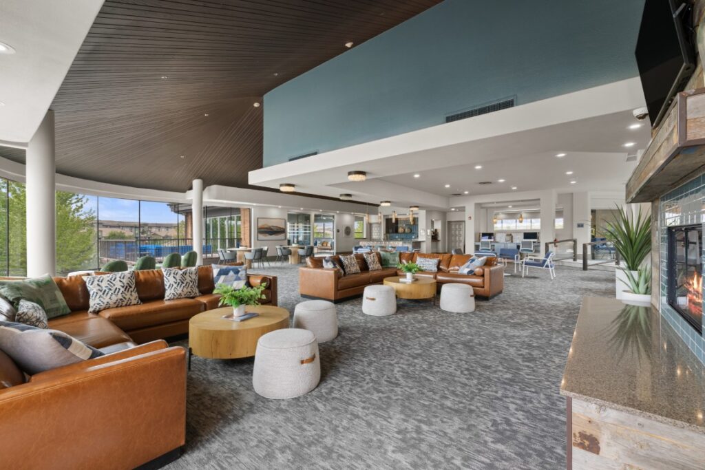 Modern lounge with brown leather sofas, round wooden tables, gray poufs, and a fireplace. Large windows let in natural light. Open kitchen and dining area are visible in the background. Contemporary decor throughout.