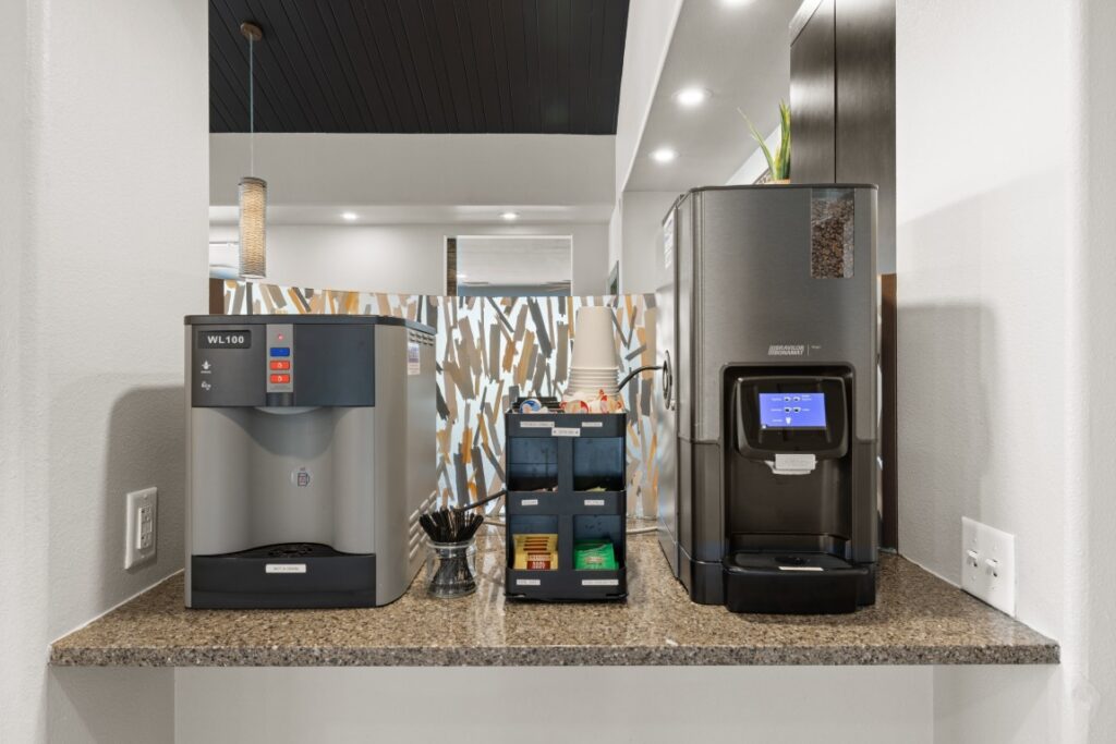 A coffee station with two coffee machines, a supply organizer holding cups, lids, stirrers, sugar packets, and creamers, all set on a granite countertop in a modern setting.