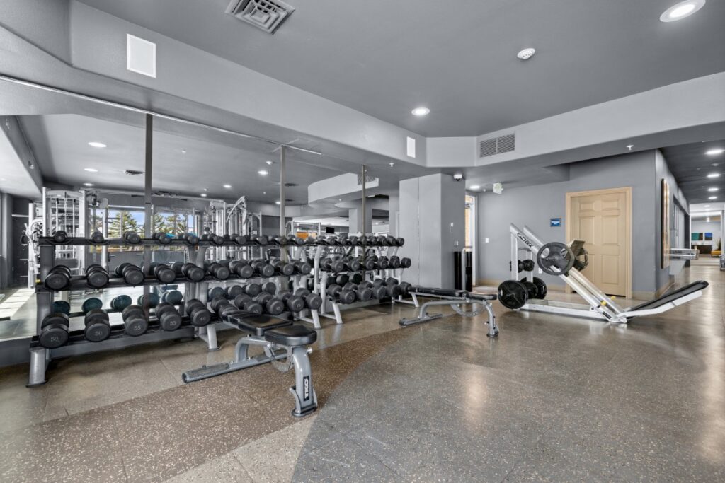 A modern gym with weight racks, dumbbells, benches, mirrors, and a leg press machine. The room has gray walls, a tiled floor, and bright lighting.