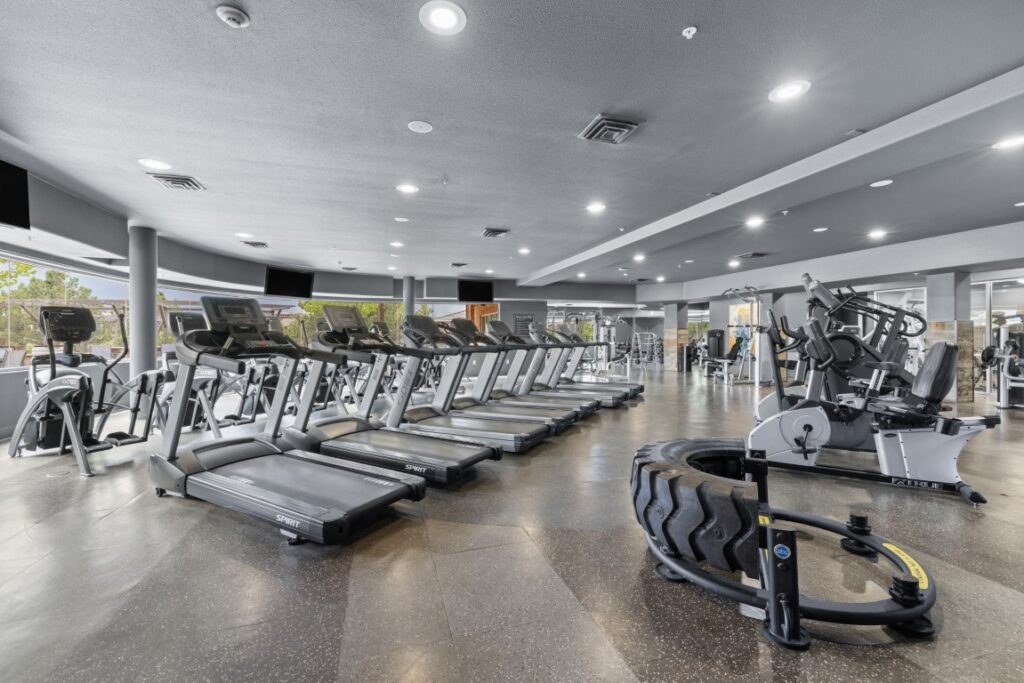 Spacious gym with several treadmills, ellipticals, stationary bikes, and large tires for functional training on a polished floor, with windows providing natural light.