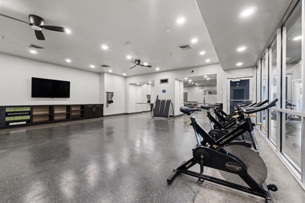 Spacious, modern gym with stationary bikes, a wall-mounted TV, ceiling fans, storage cubbies, and large windows. The room has gray flooring and bright overhead lighting.