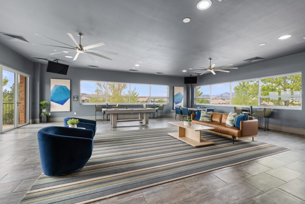 Modern lounge with large windows, two blue chairs, a tan sofa, a striped rug, a table, wall art, two pool tables, and ceiling fans. Trees and mountains are visible through the windows.