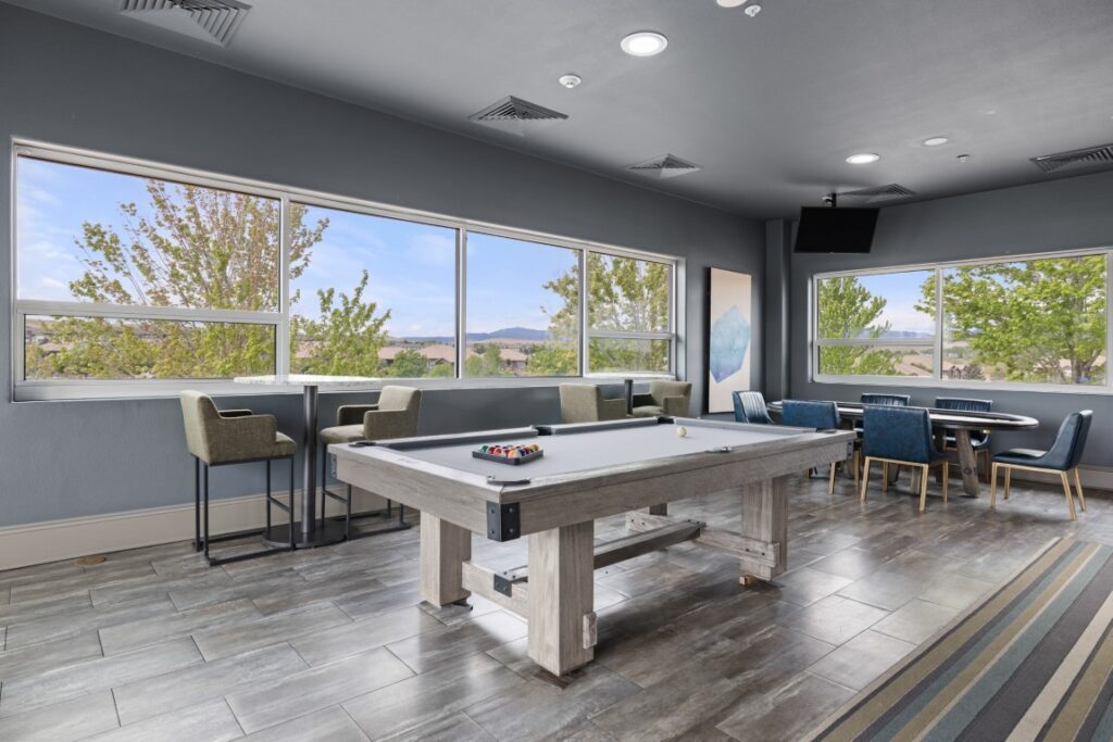 Modern game room with a white pool table, cue sticks, high chairs by large windows, a round table with chairs, and outdoor scenery visible through the windows.