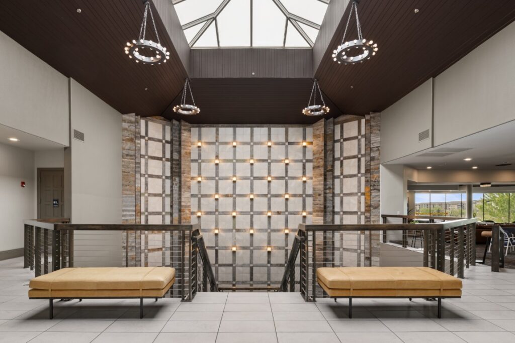 Modern hotel lobby with beige benches, a geometric lighted wall, metal railings, circular chandeliers, and a skylight above. Large windows provide natural light and a view of trees outside.
