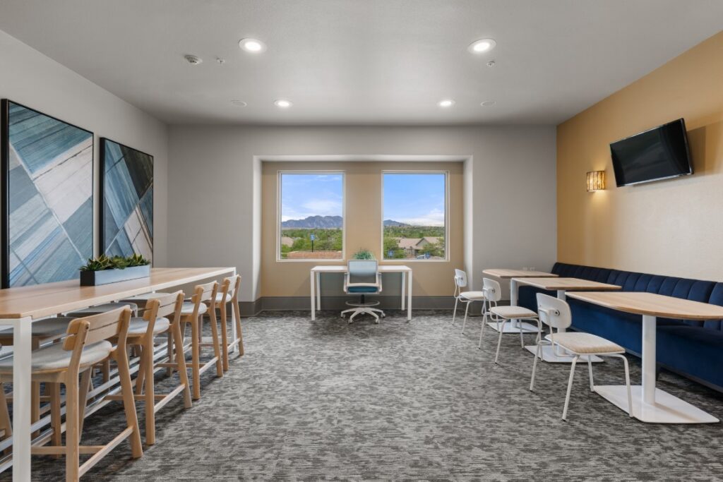 Modern lounge with a long high table and chairs, small tables with white chairs, a blue cushioned bench, wall art, mounted TV, and large windows showing a mountain view under a blue sky.