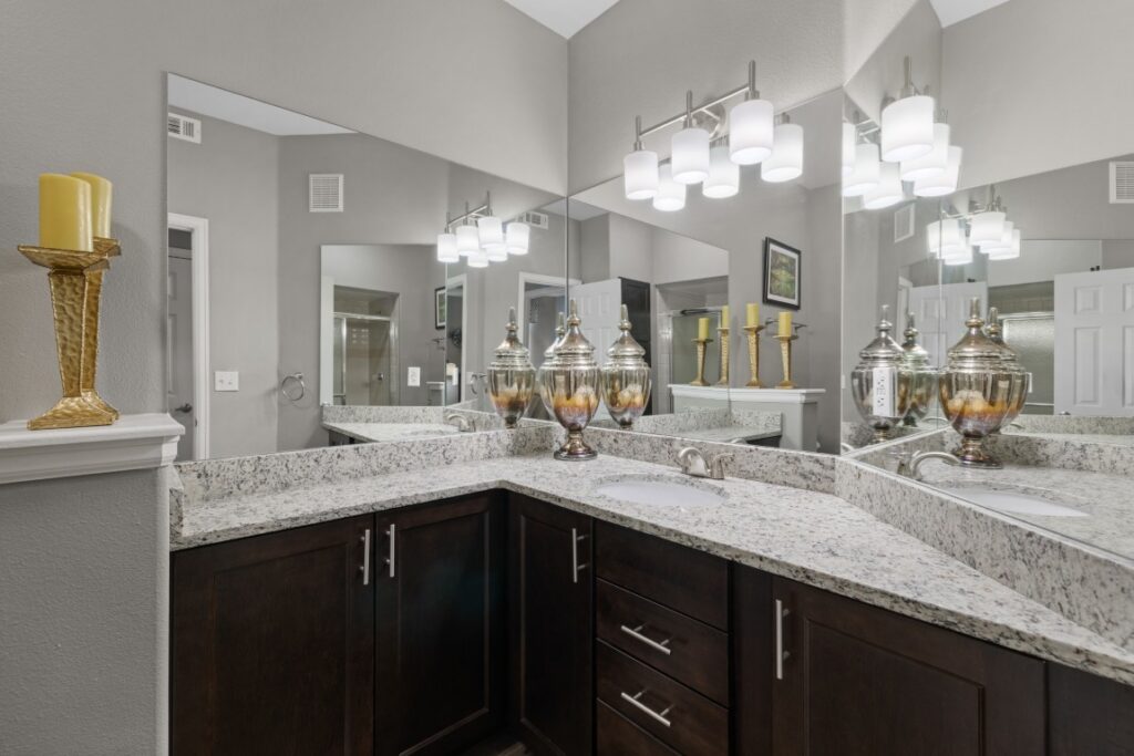 Modern bathroom with granite counters, dark cabinets, dual sinks, three decorative jars, candles on a shelf and counter, large mirrors, and contemporary lighting fixtures on a gray wall.