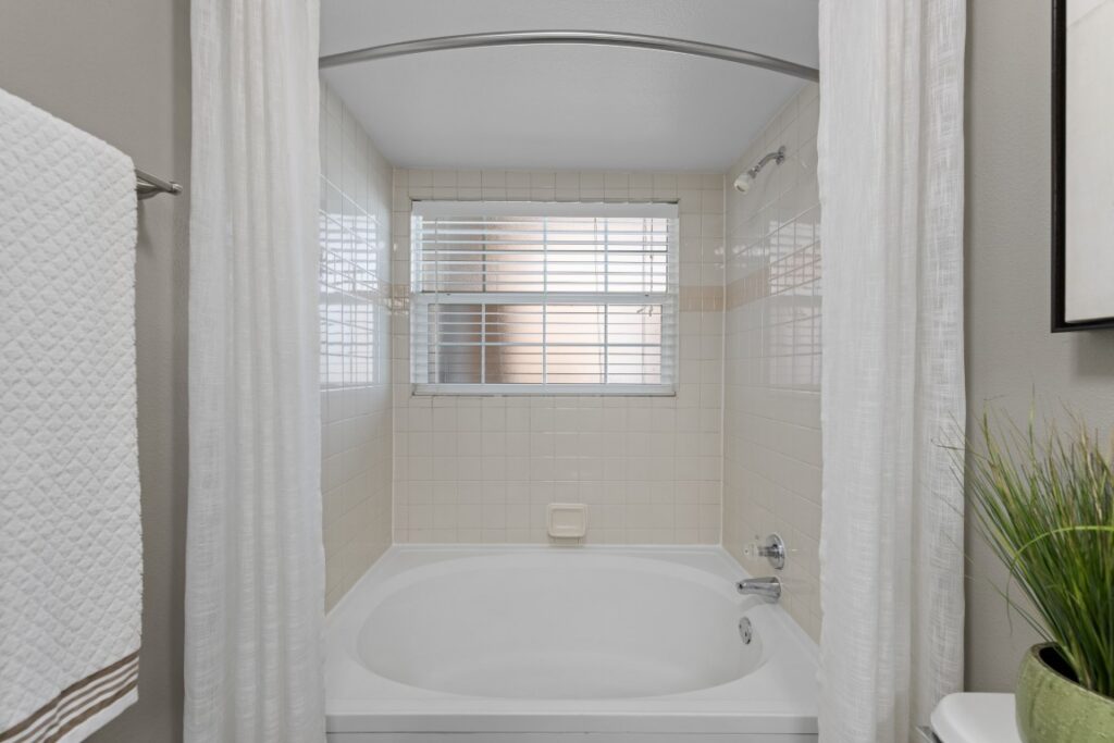White bathtub with light beige tile surround beneath a window with blinds. White shower curtains hang on both sides. A towel is on the left, and a small plant sits on the right edge. The space looks clean and modern.