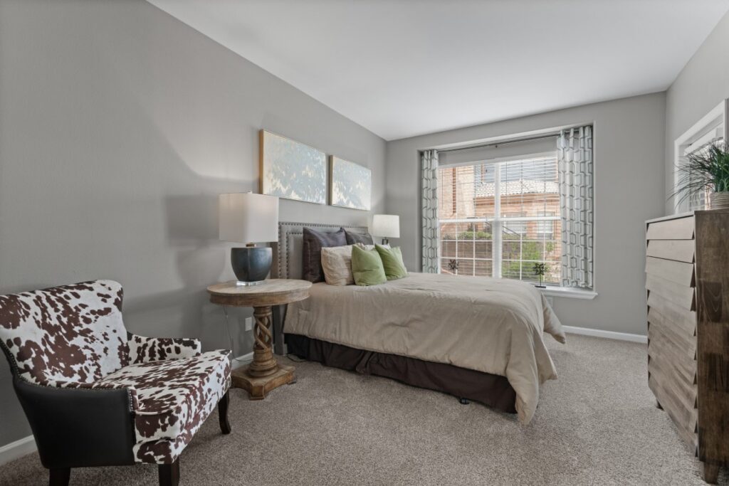 A cozy bedroom with a bed, green and beige pillows, a round wooden side table, a lamp, a dresser, a cow-print accent chair, and a large window with patterned curtains letting in natural light.
