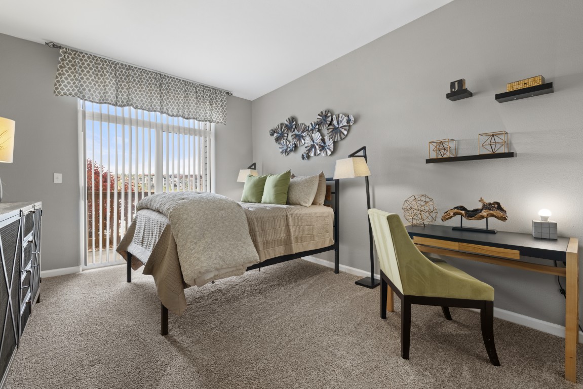 A modern bedroom with a bed, green chair at a desk, abstract wall art, shelves with decor, and a large window with vertical blinds letting in natural light.