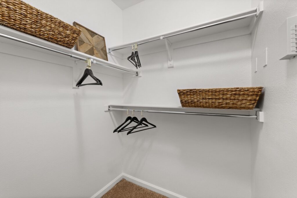Empty walk-in closet with white walls, metal shelves, black hangers, two woven baskets, and a framed wall art piece in the corner. Carpeted floor is visible at the bottom.