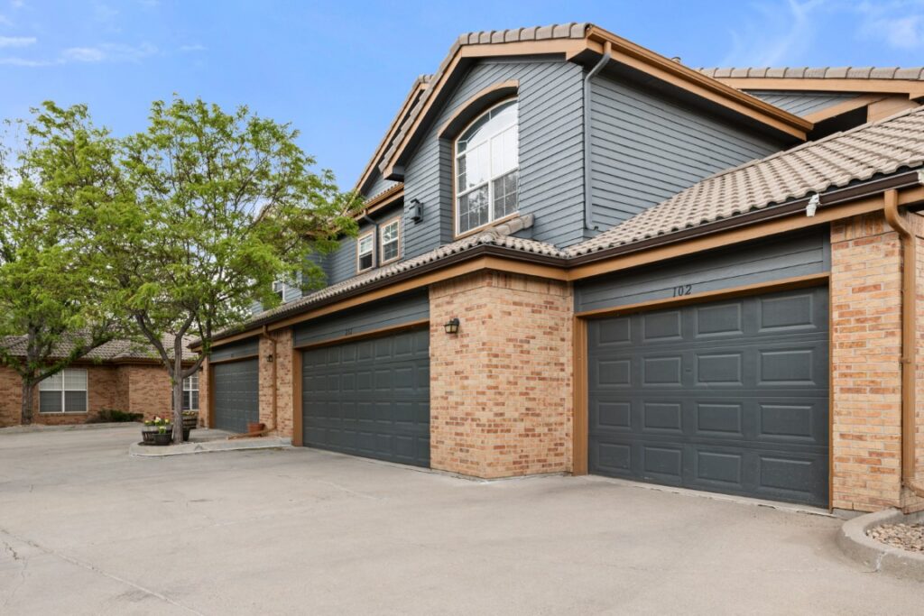Two-story townhouse with brick and gray siding, three dark gray garage doors, light brown roof tiles, large windows, and a tree on the left side. The driveway is wide and clear under a blue sky with light clouds.