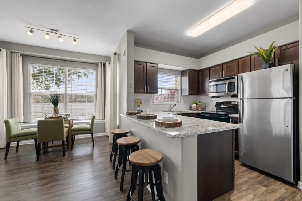 Modern kitchen with granite countertops, stainless steel appliances, dark wood cabinets, and a breakfast bar with stools. Dining area with green chairs and a window with curtains lets in natural light.