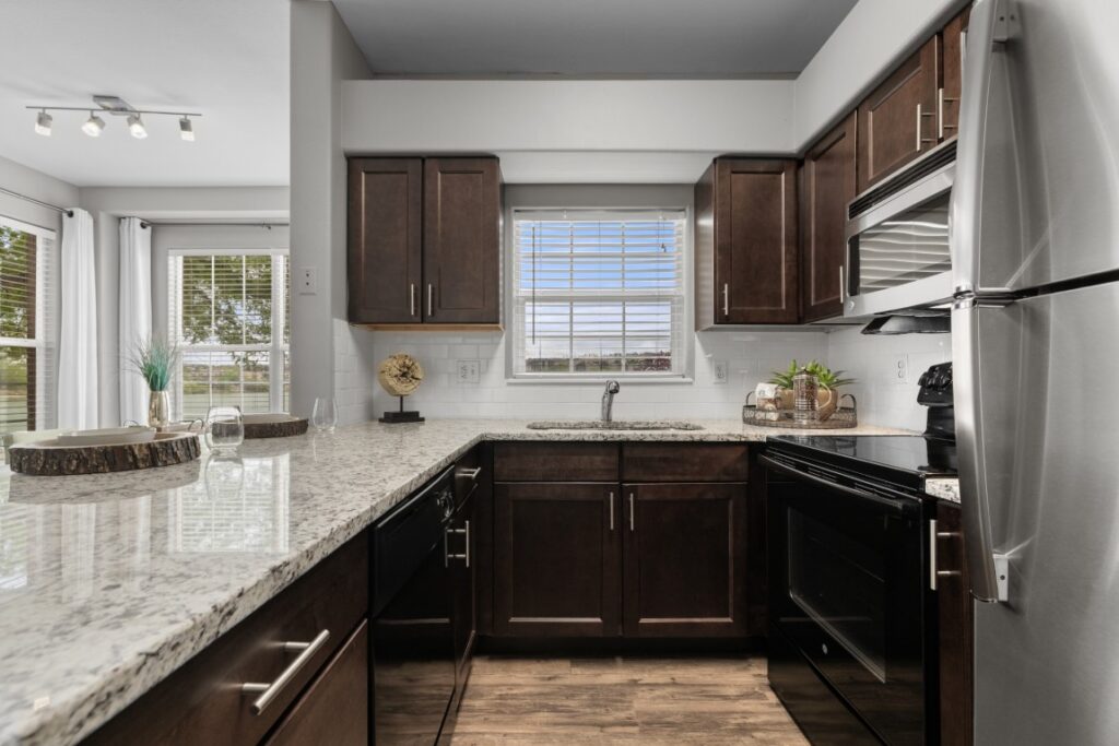 Modern kitchen with dark wood cabinets, granite countertops, stainless steel appliances, a large window above the sink, and decorative items on counters. Bright natural light fills the room from windows and glass doors.