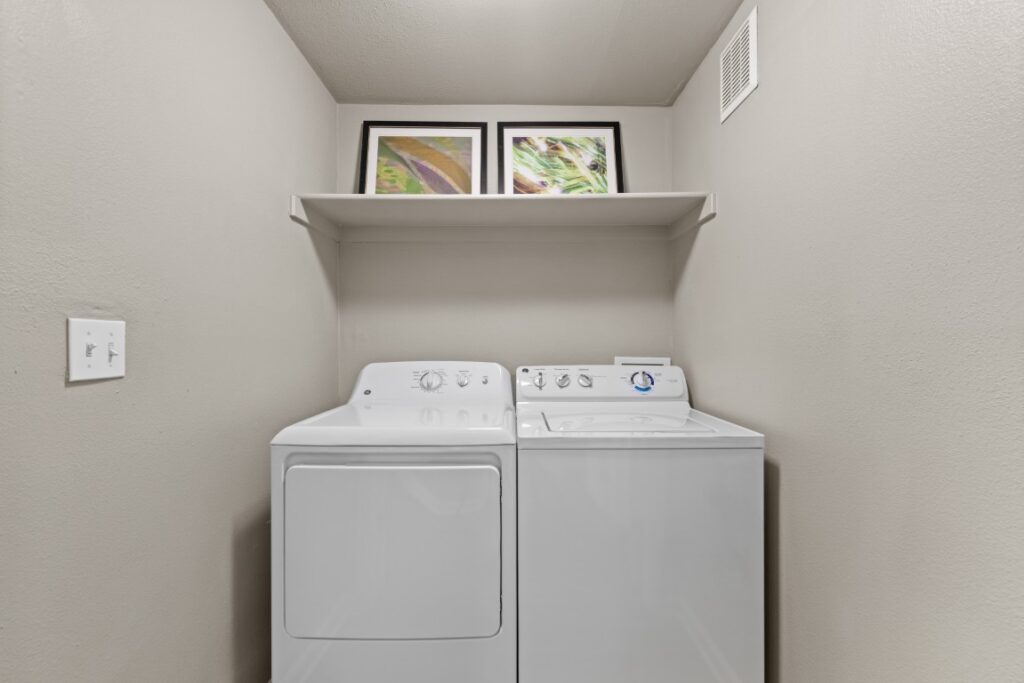 A laundry area with a white dryer and washer below a shelf holding two framed pictures, set against beige walls with a vent and a light switch visible.