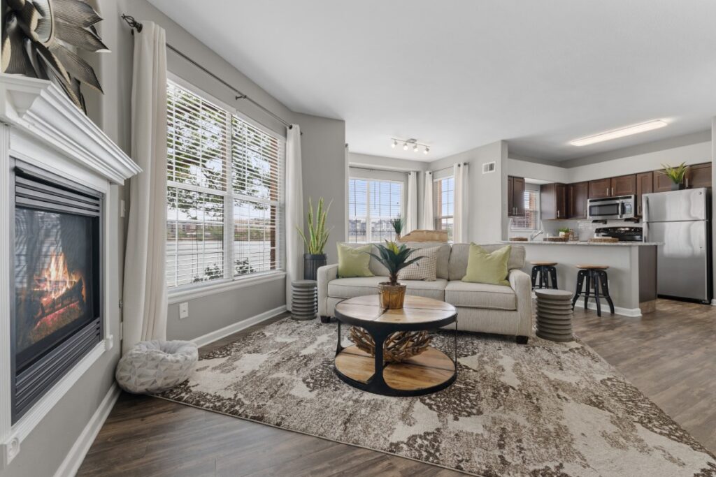 Modern living room with a fireplace, light sofa, green plants, round coffee table, patterned rug, barstools, and a kitchen in the background, all with neutral and natural tones.