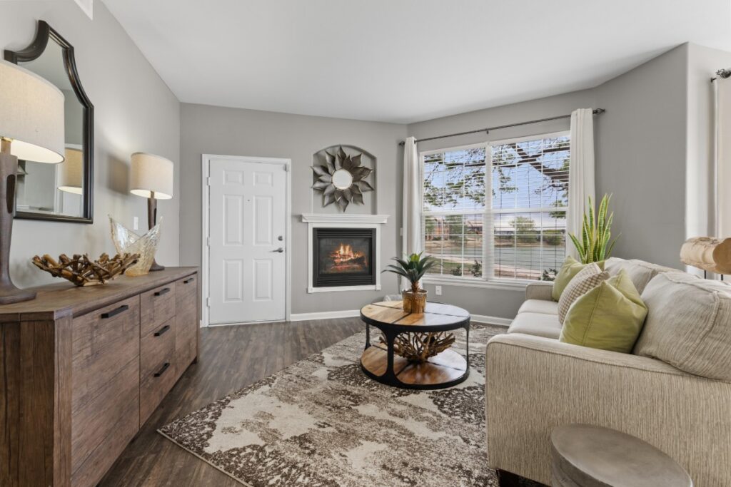 Modern living room with wood floors, a beige sofa, a round coffee table, a fireplace in the corner, and large windows with white curtains letting in natural light. Decor includes plants, lamps, and wall art.