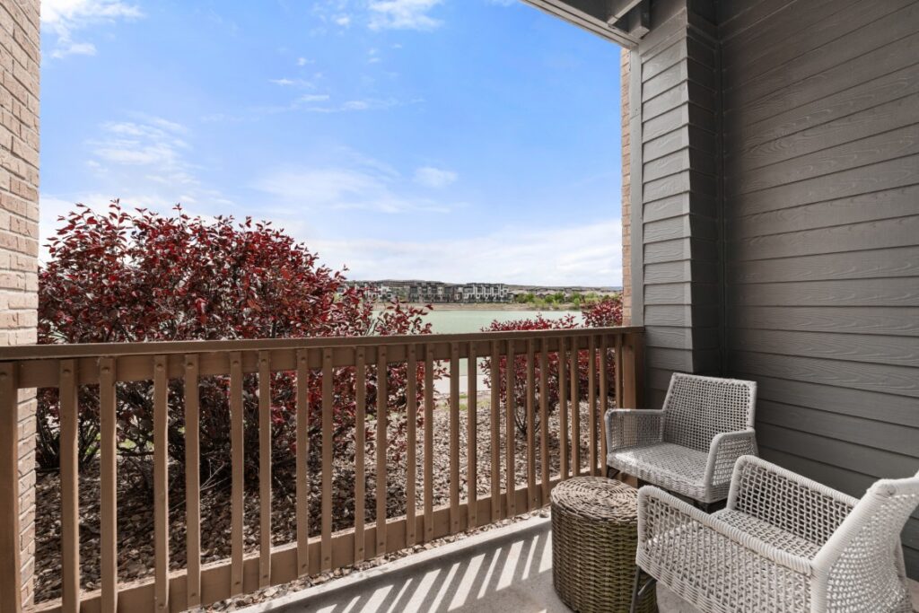 Cozy patio with two white chairs and a small table overlooks red shrubs, a wooden fence, and a serene lake under a bright blue sky with light clouds.
