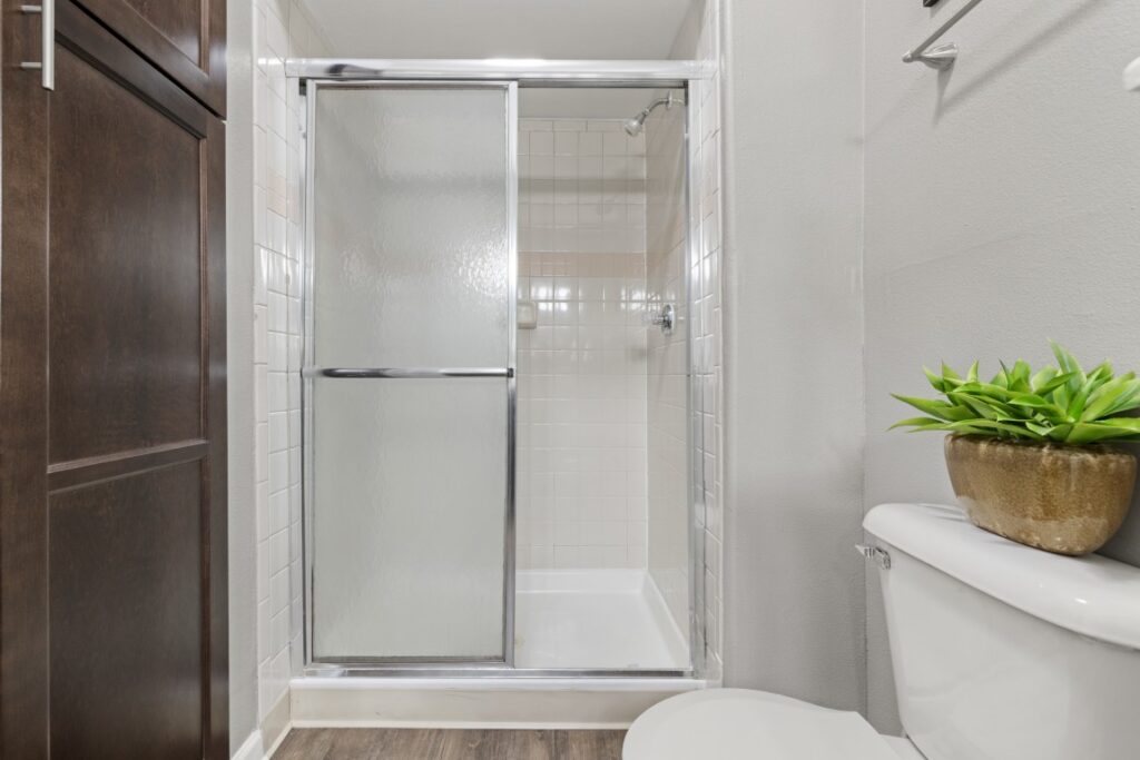 A bathroom with a glass shower door, white tiled shower, white toilet, a potted plant on the toilet tank, light walls, and wood-look flooring.