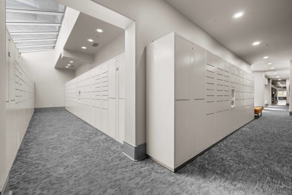 A clean, modern hallway with white lockers on both sides, gray carpet, bright lighting, and a skylight above. A seating area with chairs is visible at the end of the hall.