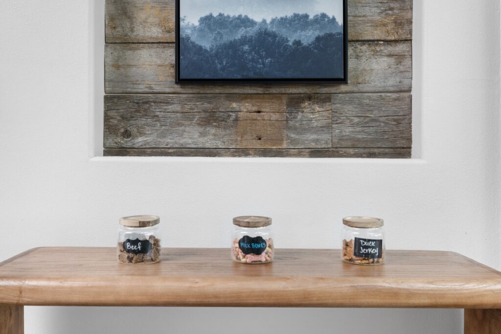 Three glass jars labeled Beef, Milk Bones, and Dixie Jerky sit on a wooden table. Behind them is a rustic wooden accent wall with a framed photo of misty mountains.