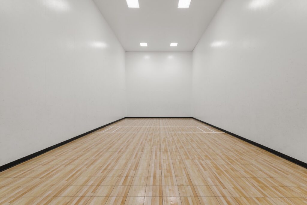 Empty indoor racquetball court with wood flooring, white walls, and bright overhead lighting. The court markings are visible on the floor.