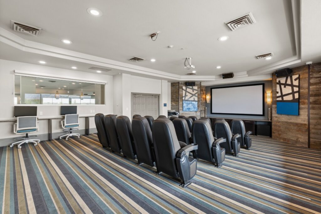 A modern home theater room with rows of black recliner seats facing a large screen, striped carpet, wood-paneled walls, and a small desk area with computers in the back.